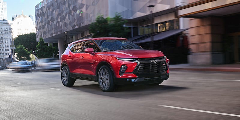 An all-new red 2024 Chevy Blazer driving through the downtown streets of a large city. - Jim Turner Chevrolet McGregor, TX