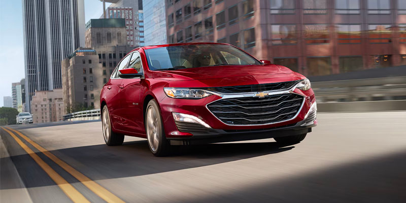 An all new red 2025 Chevy Malibu driving through downtown roads. - Jim Turner Chevrolet McGregor, TX